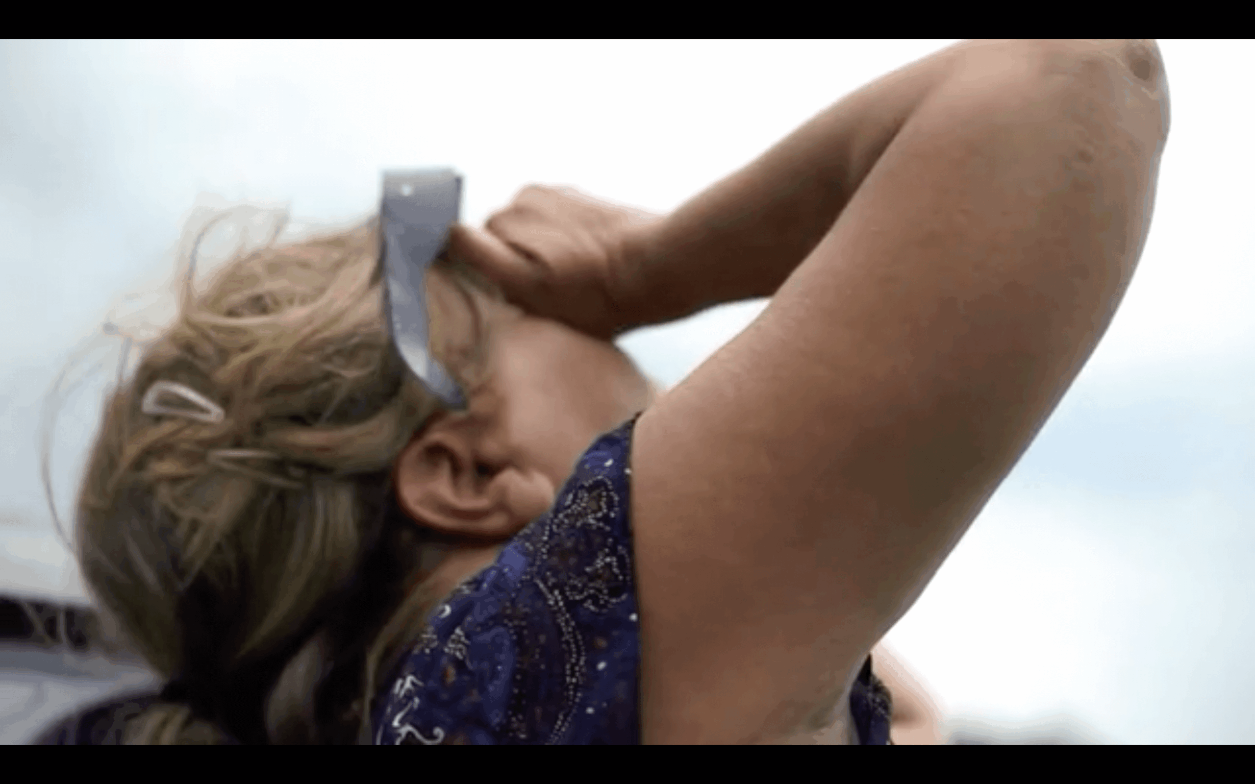 an image of a child with long bling hair looking up to the sky without glasses. they cover their eyes with the hands