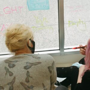 two people writing on a window, one holding a red marker