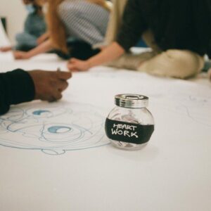 Someone drawing a face in blue on a roll of white paper. a jar with the text "Heart work" sits next to them