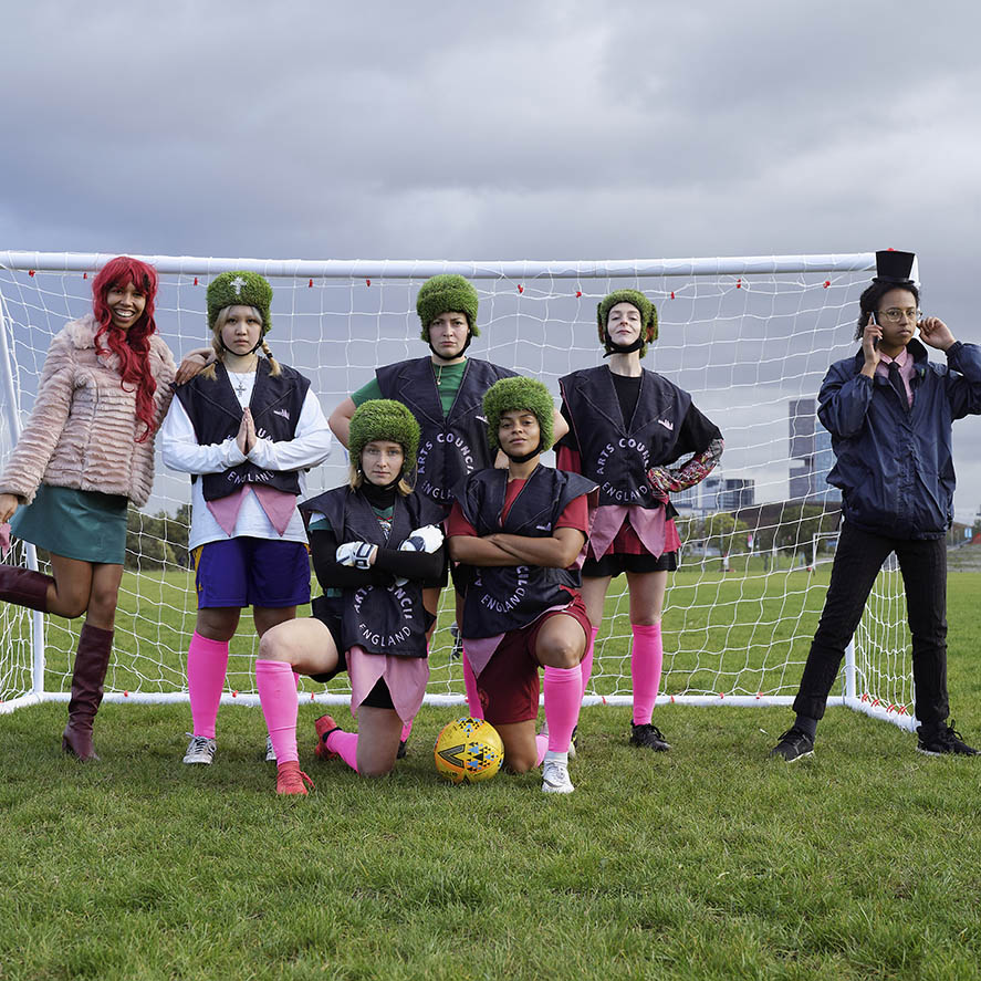 a photograph of seven people posing in front of a football goal. five are wearing sports kits with "arts council england" painted on to them and wearing helmets covered in grass.