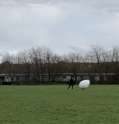 Katherine, a white, neurodivergent woman artist wearing black sports clothes is running behind a giant white egg on a green recreation ground next to a train track. She is on her own. There is a red South Western Railway train going across in the background, and below it there are low warehouses / workshops / shed-like buildings. The sky is grey and bright and the trees don’t have leaves.