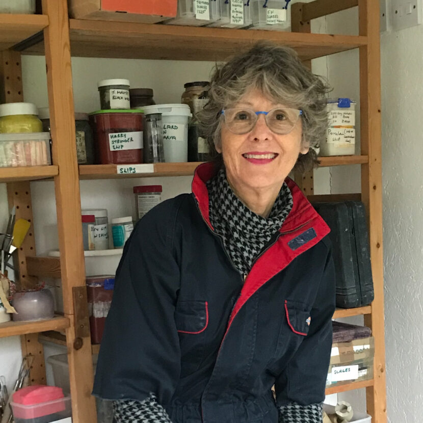 a photograph of sandy layton. sandy is standing in front of a shelf of art supplies. sandy is wearing a black and white sweater and dark blue coveralls.