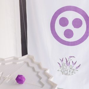 a white tapestry with a purple circle with three dots inside and an image of lavender underneath hangs in the background whilst a purple d20 sits on a white table in the foreground