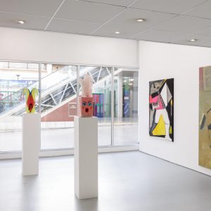 a sculpture of a jester mask and a pink box with a large feather are displayed on plinths with a painting of someone sitting and reading and a geometric painting are being displayed on the wall