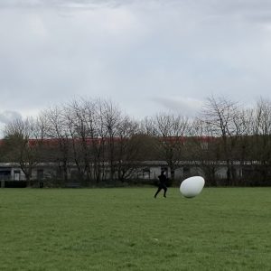Katherine, a white, neurodivergent woman artist wearing black sports clothes is running behind a giant white egg on a green recreation ground next to a train track. She is on her own. There is a red South Western Railway train going across in the background, and below it there are low warehouses / workshops / shed-like buildings. The sky is grey and bright and the trees don’t have leaves.
